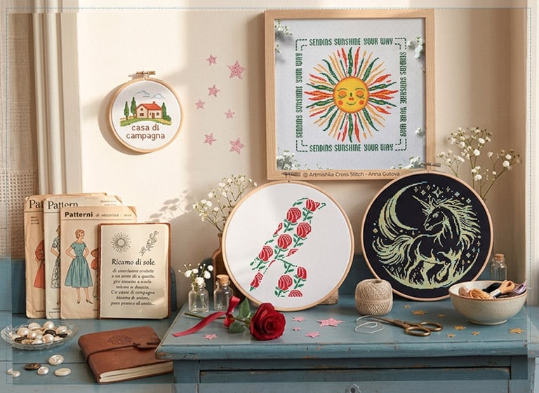 Broderies au point de croix Artmishka sur une table vintage : soleil souriant, initiale fleurie et licorne, entourées de patrons et de fleurs des champs.